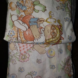 May include: Two white cotton twin-sized bed sheets with a floral pattern. The top sheet features a cartoon girl with blonde hair wearing a red dress and holding a book. The bottom sheet features a cartoon girl with blonde hair wearing a red and white checkered dress and holding a ball.