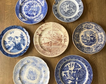 Puede incluir: Una colección de siete platos decorativos antiguos con varios diseños azules y blancos. Los platos presentan escenas de paisajes, figuras y diseños florales. Un plato es marrón y blanco con un diseño de inspiración asiática.