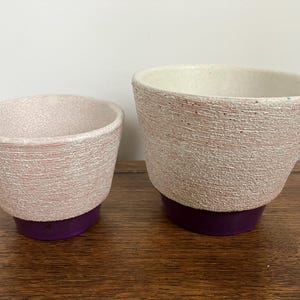 May include: Two white ceramic planters with a textured finish and a purple base. The larger planter is taller than the smaller planter.