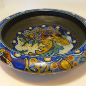Op de afbeelding: Een decoratieve keramische kom met een donker interieur en een brede, sierlijke rand. De rand heeft een blauwe basis met kleurrijke bloemen- en geometrische patronen in oranje, geel en wit. De binnenkant van de kom heeft een bloemmotief.