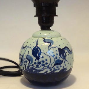 Op de afbeelding: Een keramische tafellampvoet met een zwarte lamphouder. De voet is rond met een lichtgroene achtergrond en blauwe bloemmotieven. De onderkant van de voet is donkerblauw en een zwart snoer loopt van de lamp.