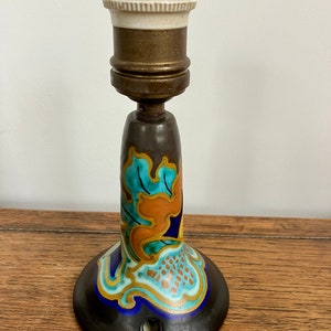 Op de afbeelding: Een keramische wandlamp met een blauw, groen, oranje en gouden bloemenmotief. De lamp heeft een witte porseleinen fitting en een messing armatuur.