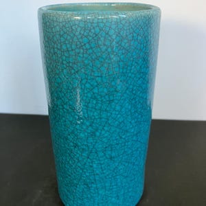 May include: A tall, cylindrical turquoise vase with a crackle glaze finish. The vase has a smooth, rounded rim and a dark, textured pattern throughout the surface. The color transitions from a lighter turquoise at the top to a deeper teal towards the base.