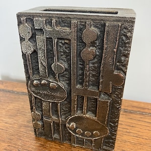 Puede incluir: Un jarrón de metal plateado con un diseño geométrico. El jarrón tiene una forma rectangular y está hecho de metal texturizado.