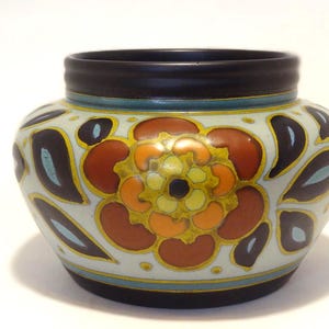 Peut inclure: Un vase en céramique décoratif avec un bord et une base marron foncé. Le vase présente un motif floral dans les tons orange, marron et jaune, sur un fond crème. Le motif comprend des accents marron foncé et turquoise. Environ 15 cm de haut.