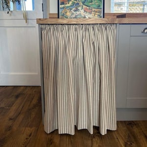 Puede incluir: Una cortina de tela rayada beige y marrón con un ribete superior fruncido, colgada delante de un armario de cocina. La cortina está hecha de una tela ligera y tiene un diseño simple y elegante.