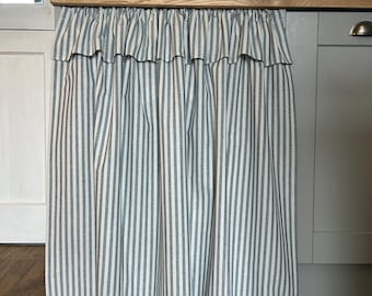May include: A kitchen sink skirt made of fabric with a blue and white vertical striped pattern. The curtain features a gathered ruffle along the top edge and hangs straight down, covering the lower cabinet area beneath a wooden countertop.