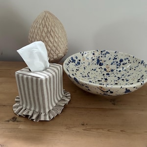 May include: A square tissue box cover featuring beige and white vertical stripes with a ruffled base, a white tissue protruding from the top, a beige textured decorative object, and a wide ceramic bowl with blue and tan paint splatters.