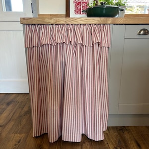 Puede incluir: Un mueble de cocina con una cortina de tela a rayas rojas y blancas. La cortina está fruncida en la parte superior y cuelga hasta el suelo. Una encimera de madera se encuentra encima de la cortina, con un estampado enmarcado y una olla verde.