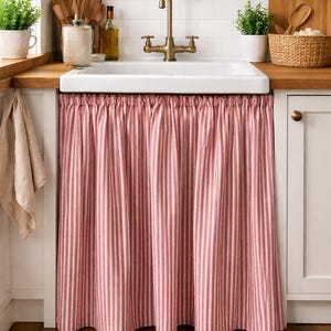 Puede incluir: Un fregadero de cocina con un lavabo blanco y un grifo de latón. Una cortina a rayas rojas y blancas cuelga debajo del fregadero. La encimera es de madera y hay plantas y utensilios de madera cerca.