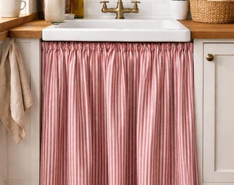 May include: A kitchen sink with a white basin and brass faucet. A red and white striped curtain hangs below the sink. The countertop is wood, and there are plants and wooden utensils nearby.