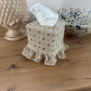 May include: A square fabric tissue box cover in a light beige colour with a small black floral pattern. The cover features a ruffled hem along the bottom edge and a square opening at the top, through which a single white facial tissue is protruding.