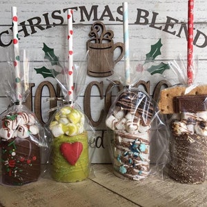 May include: Four different hot chocolate on a stick, each with a different topping. The hot chocolate is in a clear plastic bag with a white and red polka dot straw. The toppings include marshmallows, chocolate chips, sprinkles, and graham crackers. The background is a white wooden plank with the words "Christmas Blend Hot Cocoa" written on it.