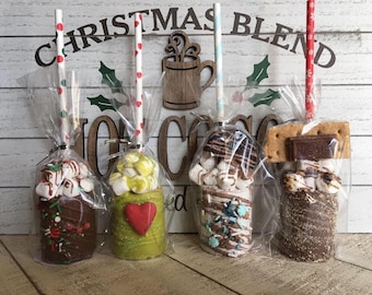 May include: Four different hot chocolate on a stick, each with a different topping. The hot chocolate is in a clear plastic bag with a white and red polka dot straw. The toppings include marshmallows, chocolate chips, sprinkles, and graham crackers. The background is a white wooden plank with the words "Christmas Blend Hot Cocoa" written on it.