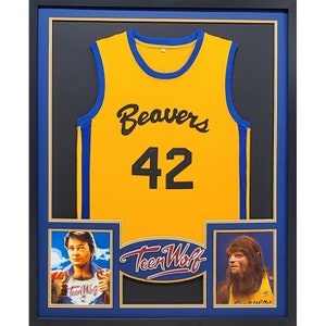May include: A framed yellow basketball jersey with blue trim, featuring the text "Beavers" and the number "42". The jersey is displayed with two images, one of a man in a red jacket and the other of a man with a furry face. The text "Teen Wolf" is displayed in a blue and gold banner.
