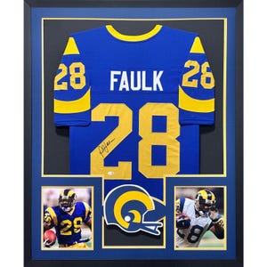 May include: A framed sports display featuring a blue and yellow jersey with the name FAULK and number 28, signed by the athlete. Below are two photographs of a player in a matching uniform, flanking a Rams team logo, all set in a black frame.