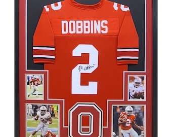 May include: A framed red and white Ohio State football jersey with the name "DOBBINS" and the number "2" on the back. The jersey is signed by the player. The frame includes four action photos of the player in a game.