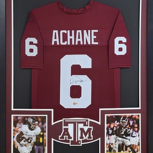 May include: A framed maroon jersey with the name "ACHANE" and the number "6" on the back. The jersey is signed with a white signature. The frame includes two smaller photos of a football player wearing the same jersey.