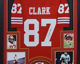 Könnte beinhalten: Ein gerahmtes rotes Footballtrikot mit der Nummer 87 und dem Namen "CLARK" auf der Rückseite. Das Trikot ist von "Dwight Clark" signiert. Der Rahmen enthält vier kleinere Fotos von Clark beim Footballspielen.