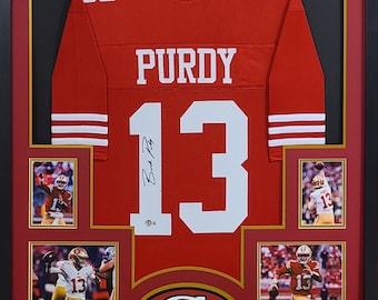 Könnte beinhalten: Ein gerahmtes rotes Footballtrikot mit dem Namen "PURDY" und der Nummer "13" in Weiß. Das Trikot ist von "Brock Purdy" signiert. Der Rahmen enthält drei kleinere Fotos des Spielers in Aktion.