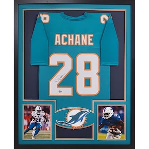 May include: A framed Miami Dolphins jersey with the number 28 and the name "Achane" on the back. The jersey is teal blue with white and orange accents. The frame is black and includes a small photo of a dolphin and two photos of players in action.