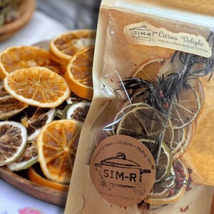 May include: A clear bag of dried citrus slices, rosemary, and berries labelled "SIM-R Citrus Delight Handcrafted Simmer Pot Mix." The bag is partially open. A wooden bowl holds additional dried citrus slices. The product is for simmer pots.