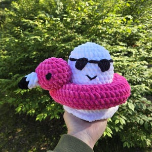 Könnte beinhalten: Ein handgefertigtes Häkelflamingo-Spielzeug mit weißem Körper, rosa Hals und rosa Schwimmring. Der Flamingo trägt eine schwarze Sonnenbrille und hat ein lächelndes Gesicht. Das Spielzeug wird vor einem grünen, belaubten Hintergrund gehalten.