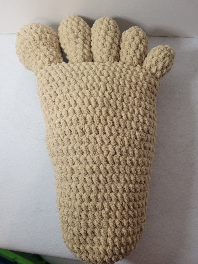 May include: A beige knitted foot-shaped pillow. The pillow is made of a soft, fluffy yarn and has five toes.