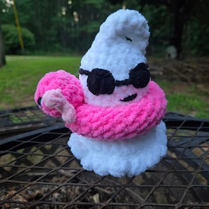 May include: A white crocheted ghost plushie wearing black yarn sunglasses and a bright pink crocheted pool floatie around its middle. The ghost has a soft, fuzzy texture and a small black stitched mouth, appearing as a decorative stuffed toy figure.
