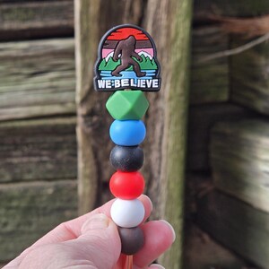 May include: A Bigfoot-themed straw topper with a colorful design. The topper features a cartoon image of Bigfoot with the text "WE BELIEVE". The straw topper has a series of colorful beads in green, blue, black, red, white, and brown.