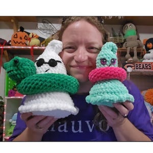 May include: Two crochet plushies: a white ghost wearing black sunglasses with a green snake wrapped around it, and a light green cone-shaped plushie with large pink and black eyes wearing a bright pink ring around its middle.