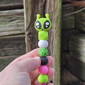 May include: A beaded alien-themed pen with a green alien head, a green cube, white, green paisley, black, pink, and lime green beads. The pen has a pink stem.