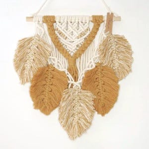 May include: A white and mustard yellow macrame wall hanging with a geometric pattern and five feather-shaped tassels. The hanging is attached to a wooden dowel with a small brown tag that says "Zest".