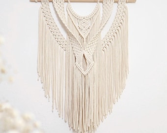 Puede incluir: Un tapiz de macramé blanco con un diseño geométrico. El tapiz está hecho de cordón de algodón y tiene una barra de madera en la parte superior.