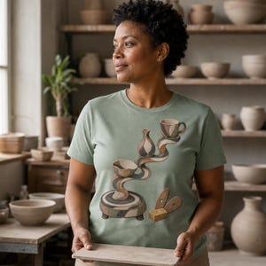 Peut inclure: Un t-shirt vert sauge à manches courtes orné d'une illustration graphique représentant une sculpture en poterie empilée et tourbillonnante avec un bol, un vase et une tasse, ainsi qu'une petite éponge rectangulaire et deux outils en bois.
