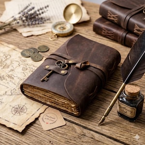 May include: A brown leather journal with a key and clasp closure, vintage style. The journal is surrounded by a compass, coins, a feather quill, and a small bottle of ink. A map and a tag that says "Handmade with Love" are also present.