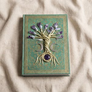 May include: A green hardcover book featuring a gold-toned wire tree sculpture on the front cover. The tree branches are tipped with purple amethyst crystals, and a single round purple stone sits at the base. Two silver crescent moons flank the trunk.