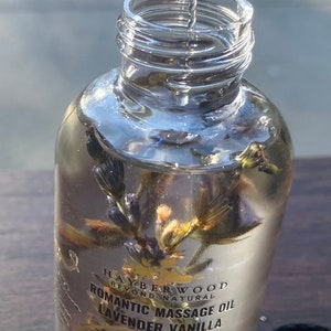 Pu&ograve; includere: Una bottiglia di vetro trasparente con un'etichetta dorata che recita "HAYBERWOOD BEYOND NATURAL ROMANTIC MASSAGE OIL LAVENDER VANILLA". La bottiglia contiene un liquido trasparente con fiori di lavanda secchi all'interno. La bottiglia sta per essere riempita con un contagocce.