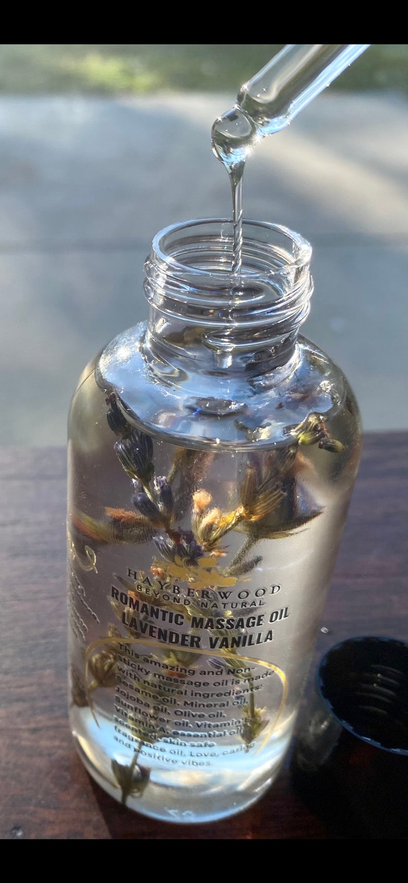 Pu&ograve; includere: Una bottiglia di vetro trasparente con un'etichetta dorata che recita "HAYBERWOOD BEYOND NATURAL ROMANTIC MASSAGE OIL LAVENDER VANILLA". La bottiglia contiene un liquido trasparente con fiori di lavanda secchi all'interno. La bottiglia sta per essere riempita con un contagocce.