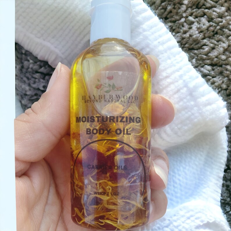 May include: A clear glass bottle with a white cap filled with yellow oil and dried flowers. The bottle label reads "Hayberwood Beyond Natural LLC Moisturizing Body Oil Carrier Oils Weight 1 oz."