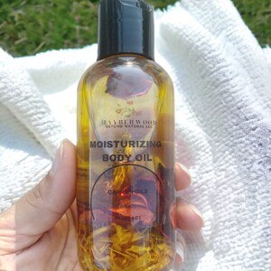 May include: A clear glass bottle with a black cap filled with a yellow liquid and dried rose petals. The label reads "Moisturizing Body Oil" and "Carrier Oils".