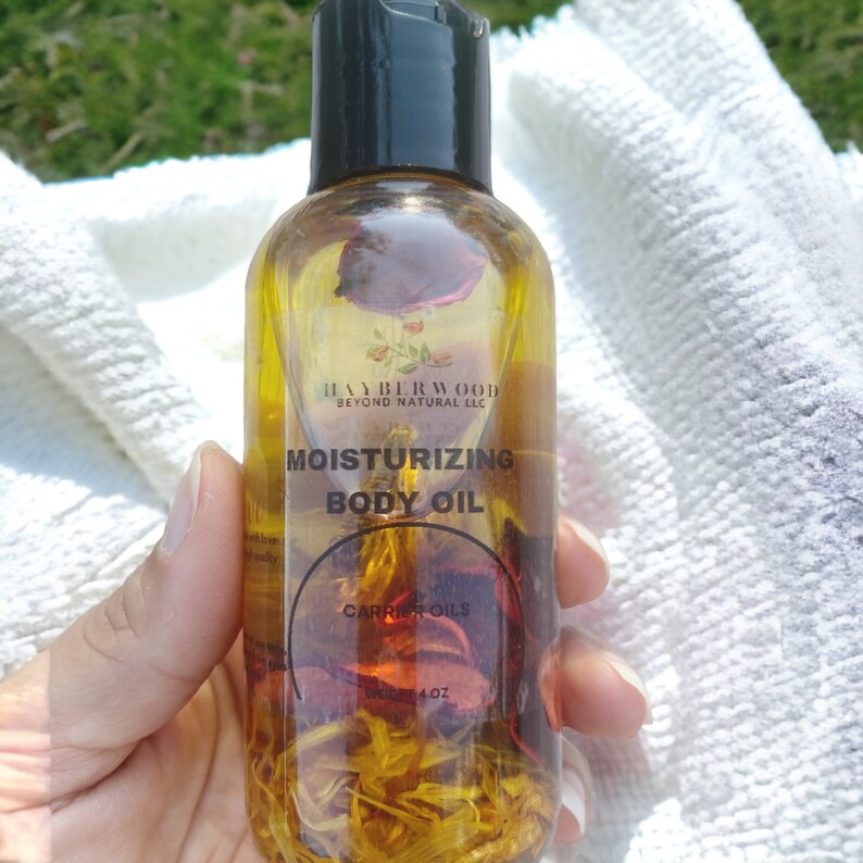 May include: A clear glass bottle with a black cap filled with a yellow liquid and dried rose petals. The label reads "Moisturizing Body Oil" and "Carrier Oils".