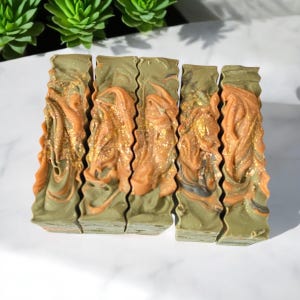 May include: Five bars of handmade soap with a green, brown, and gold swirl pattern. The soap is arranged in a row on a white surface.
