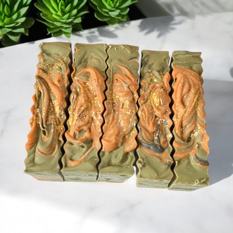 May include: Five bars of handmade soap with a green, brown, and gold swirl pattern. The soap is arranged in a row on a white surface.