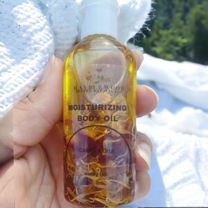 May include: A clear glass bottle with a white cap filled with a yellow liquid and dried flowers. The bottle label reads "HAYBERWOOD BEYOND NATURAL LLC" and "MOISTURIZING BODY OIL" with the text "CARRIER OILS" and "WEIGHT 1 OZ".