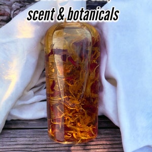 May include: A clear glass bottle filled with a golden liquid and red and yellow botanicals. The bottle is sitting on a wooden surface with a white cloth draped over it. The text "Choose the oils, scent & botanicals" is written in black text at the top of the image.