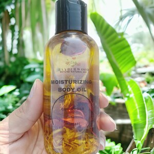 May include: A clear glass bottle with a black cap filled with a yellow liquid and dried flowers. The bottle label reads "Moisturizing Body Oil" and "Carrier Oils".
