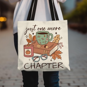 Op de afbeelding: Een witte tote bag met zwarte handvatten met een handgetekend ontwerp van een boek, een kopje koffie, een kaars en een bril. De tekst op de tas is "just one more chapter".