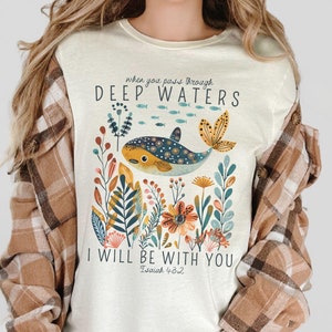 Könnte beinhalten: Ein weißes T-Shirt mit einer bunten Illustration eines Wals, der in einem Blumenfeld schwimmt. Der Text "When you pass through deep waters I will be with you Isaiah 43:2" ist auf das Shirt gedruckt.