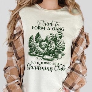 Może przedstawiać: Biały T-shirt z zielonym nadrukiem przedstawiającym trzy kury trzymające narzędzia ogrodnicze. Tekst na koszulce brzmi: "Próbowałem stworzyć gang, ale zamienił się w klub ogrodniczy".